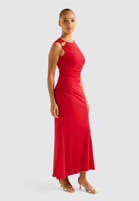 Red sleeveless dress with ruched detailing, a fitted silhouette, and side slit. Features a metal accent on the shoulder. Smooth fabric.