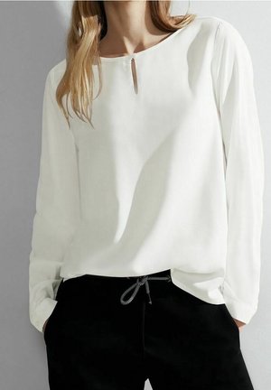 Kobieta ubrana w luźną, białą bluzkę z długim rękawem, z małym kluczykowym wycięciem przy dekolcie, oraz czarne spodnie ze sznurkiem w pasie, z rękami w kieszeniach.