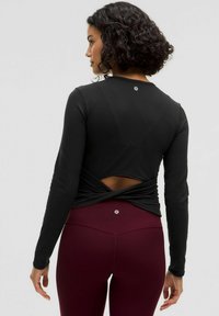Črn zgornji del z dolgimi rokavi z zasukanim dizajnom na hrbtu, v kombinaciji z maroon legicami. Ima gladko teksturo in logo na hrbtu vratu.