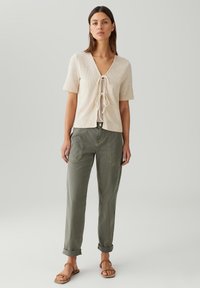 Beige gebreide top met korte mouwen, V-hals en een strikdetail aan de voorkant, gecombineerd met opgerolde olijfgroene broeken en bruine sandalen.