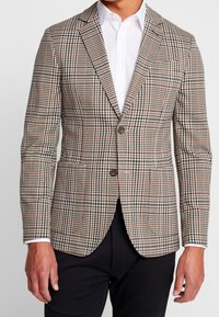 Marynarka w wzór houndstooth w odcieniach beżu, czerni i pomarańczu. Dwa guziki, klapy wycięte i kieszeń na piersi. Noszona na białej koszuli.