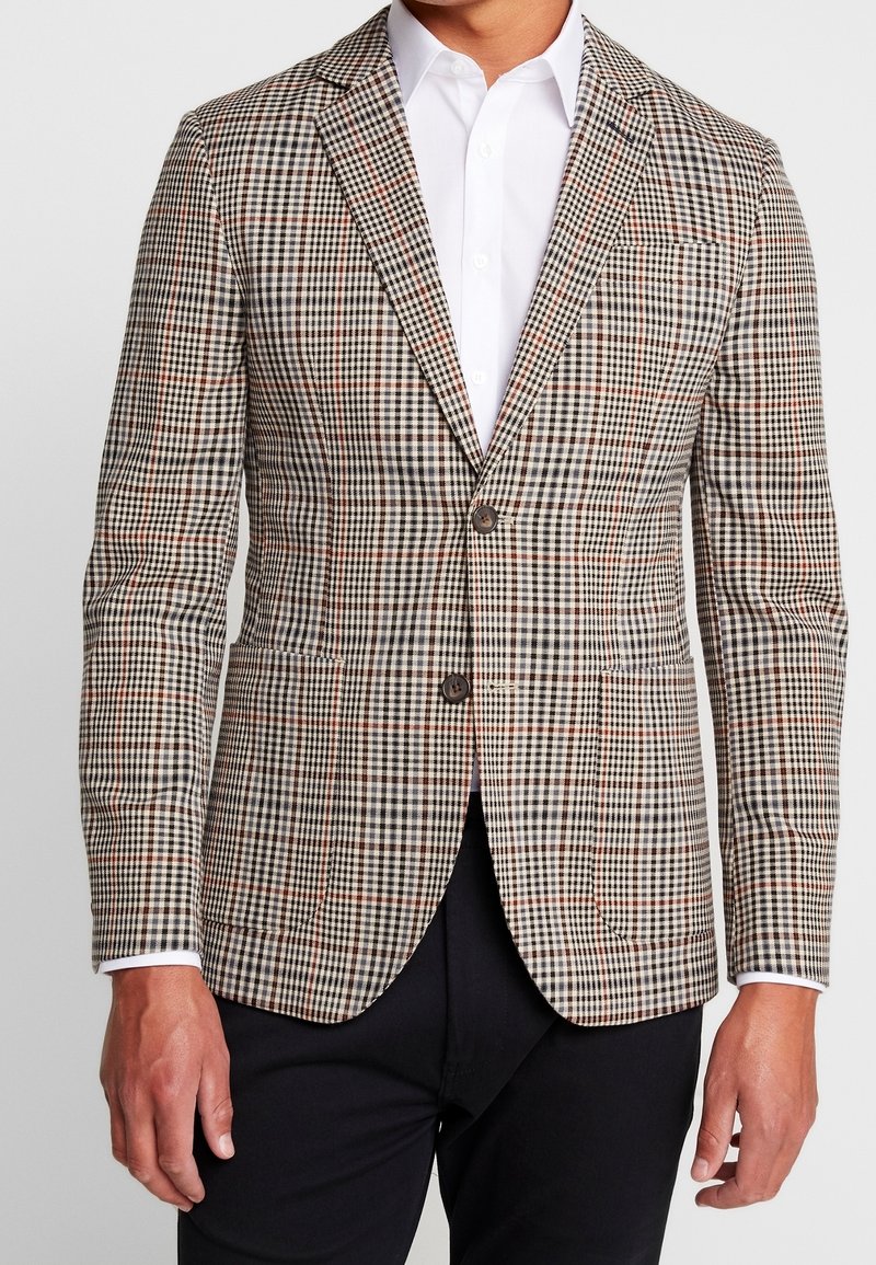 Marynarka w wzór houndstooth w odcieniach beżu, czerni i pomarańczu. Dwa guziki, klapy wycięte i kieszeń na piersi. Noszona na białej koszuli.