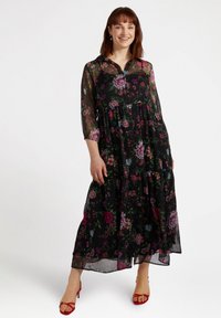 Czarna maks dress w kwiaty z przezroczystymi długimi rękawami, zaprojektowana w warstwy, ozdobiona kolorowymi kwiatami w różowych, fioletowych i niebieskich akcentach; zestawiona z czerwonymi sandałami.