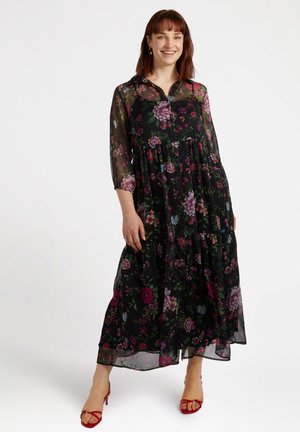 Schwarzes Maxikleid mit floralen Mustern und durchsichtigen langen Ärmeln, gestuftes Design mit bunten Blumen in rosa, lila und blauen Akzenten; kombiniert mit roten Sandalen.