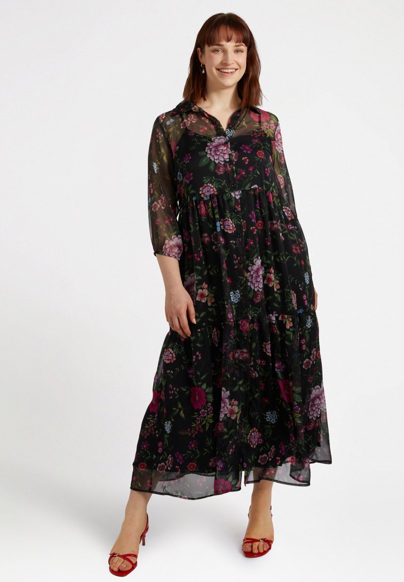 Czarna maks dress w kwiaty z przezroczystymi długimi rękawami, zaprojektowana w warstwy, ozdobiona kolorowymi kwiatami w różowych, fioletowych i niebieskich akcentach; zestawiona z czerwonymi sandałami.