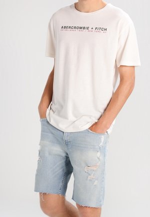 Camiseta de manga corta color crema para hombre con logo negro y rojo, combinada con shorts de mezclilla claros y desgastados con bordes deshilachados.