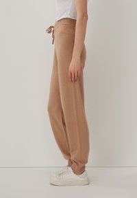 Beige, gestrickte Jogginghosen mit einem Kordelzugbund. Lockere Passform mit schmal zulaufenden Bündchen. Kombiniert mit weißen Sneakers, vor einem schlichten Hintergrund stehend.