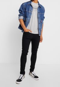 Chaqueta vaquera, camiseta gráfica gris, jeans negros ajustados y zapatillas con estampado en blanco y negro. Conjunto informal con texturas en capas.