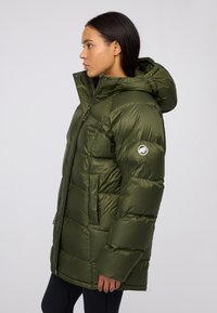 Giacca a vento verde oliva con zip frontale, cappuccio e patch con logo sulla manica sinistra. Presenta sezioni trapuntate e tasche laterali.