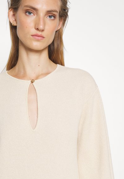 Εκρού ριχτή πλεκτή μπλούζα με λαιμόκοψη τύπου κλειδώματος και χρυσή λεπτομέρεια. Μακριά μανίκια με άνετη εφαρμογή και απαλό ύφασμα.