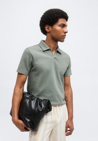 Άνδρας που φοράει πράσινο υφασμάτινο κοντομάνικο πόλο και ριγέ ανοιχτόχρωμο παντελόνι, κρατώντας μία μεγάλη μαύρη μαλακή δερμάτινη τσάντα φάκελο στο πλάι.