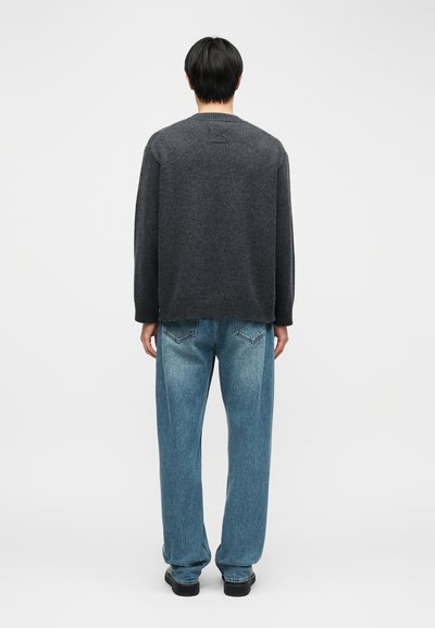 Personne aux cheveux noirs courts portant un pull gris foncé et un jean bleu, debout de dos sur un fond clair uni.