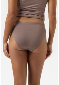 Frau, die mauvefarbene nahtlose hochgeschnittene Slips und ein passendes kurzes Oberteil trägt, steht mit entspannten Händen an ihren Seiten vor einem einfarbigen Hintergrund.