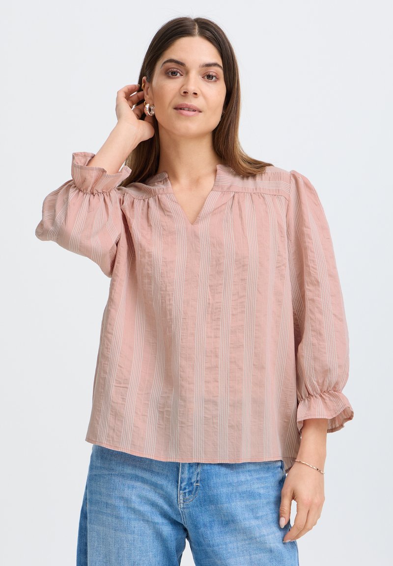 Femme aux cheveux bruns lisses portant un chemisier rose à rayures avec des manches bouffantes et un jean bleu, ajustant ses cheveux d'une main.