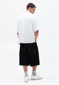 Chemise blanche à manches courtes, coupe décontractée. Short en denim noir avec deux grandes poches arrière. Des baskets blanches et des chaussettes grises complètent la tenue.