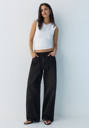Tânără care stă cu mâinile în buzunare, purtând un top alb fără mâneci, pantaloni largi negri și sandale negre, pe un fundal deschis.