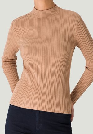 Kobieta ubrana w dopasowaną, beżową, prążkowaną bluzkę z długim rękawem, zestawioną z ciemnoniebieskimi spodniami na jednolitym tle.