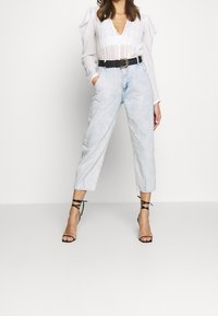 Blouse blanche à manches bouffantes, col en V et boutons à l'avant, associée à un jean court en denim bleu clair, taille haute, et des talons noirs à fines brides.