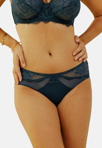 Femme portant un soutien-gorge en dentelle bleu foncé et des culottes assorties, avec des bracelets en or à son poignet gauche, les mains reposant sur les hanches.