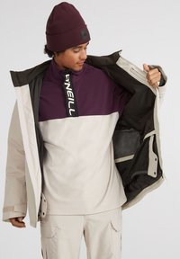 Ung person iført beige cargo-bukser og en beanie, som viser indersiden af en åben beige jakke over en lilla og beige fleece-trøje.