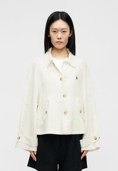 Mladá osoba nosi voľnú bielu zapínateľnú bundu s vreckami a čierne šortky na jednoliatom svetlom pozadí.