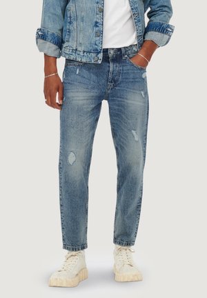 Personne portant une veste en denim bleu clair et un jean bleu déchiré avec des baskets blanches, une main dans la poche, debout devant un fond uni.