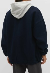 Osoba w ciemnoniebieskiej kurtce z jasnoszarym kapturem pod spodem, stojąca z rękami swobodnie wzdłuż ciała, odwrócona w stronę.