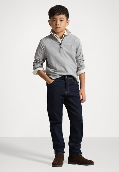 Pullover cinza com fecho, camisa branca de gola por baixo, jeans escuros, e botas de camurça castanhas até ao tornozelo; pose em pé, mãos nos bolsos.