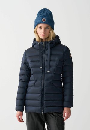 Marine blauwe pufferjack met horizontale stiksels, een rits aan de voorkant, een verstelbare capuchon en twee zijzakken; gecombineerd met een blauwe geribbelde gebreide beanie.