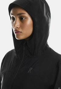 Czarna kurtka z kapturem wykonana z lekkiego, matowego materiału. Ma pełny zamek błyskawiczny, minimalne przeszycia i subtelne logo na klatce piersiowej.