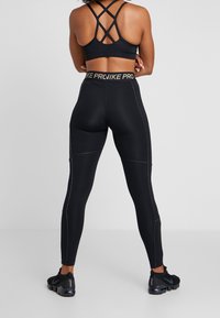 Kobieta odwrócona plecami, ubrana w czarne legginsy Nike Pro oraz czarny sportowy stanik na ramiączkach, w czarnych butach Nike, na jednolitym tle.