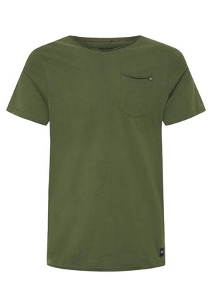 Jeune homme debout portant un t-shirt vert olive à manches courtes, un jean gris foncé et des baskets noires avec des rayures blanches, sur un fond clair uni.