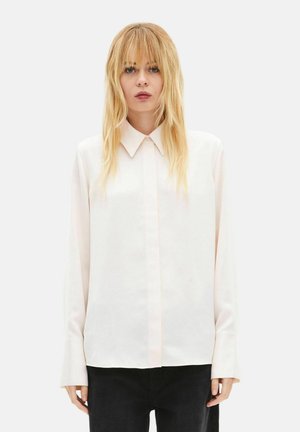 Blondynka o prostych włosach ubrana w jasnoróżową koszulę na guziki z długim rękawem i czarne spodnie, stoi na tle jednolitego tła.