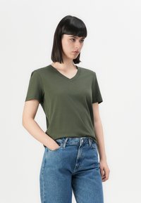 T-shirt verde oliva a scollo a V in tessuto morbido, maniche corte, abbinata a jeans blu a vita alta, esposta su uno sfondo bianco semplice.