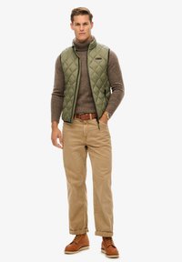 Groene gewatteerde vest over een bruine coltrui, gecombineerd met beige broeken en bruine veterschoenen, met een tan kleurige riem en een horloge.