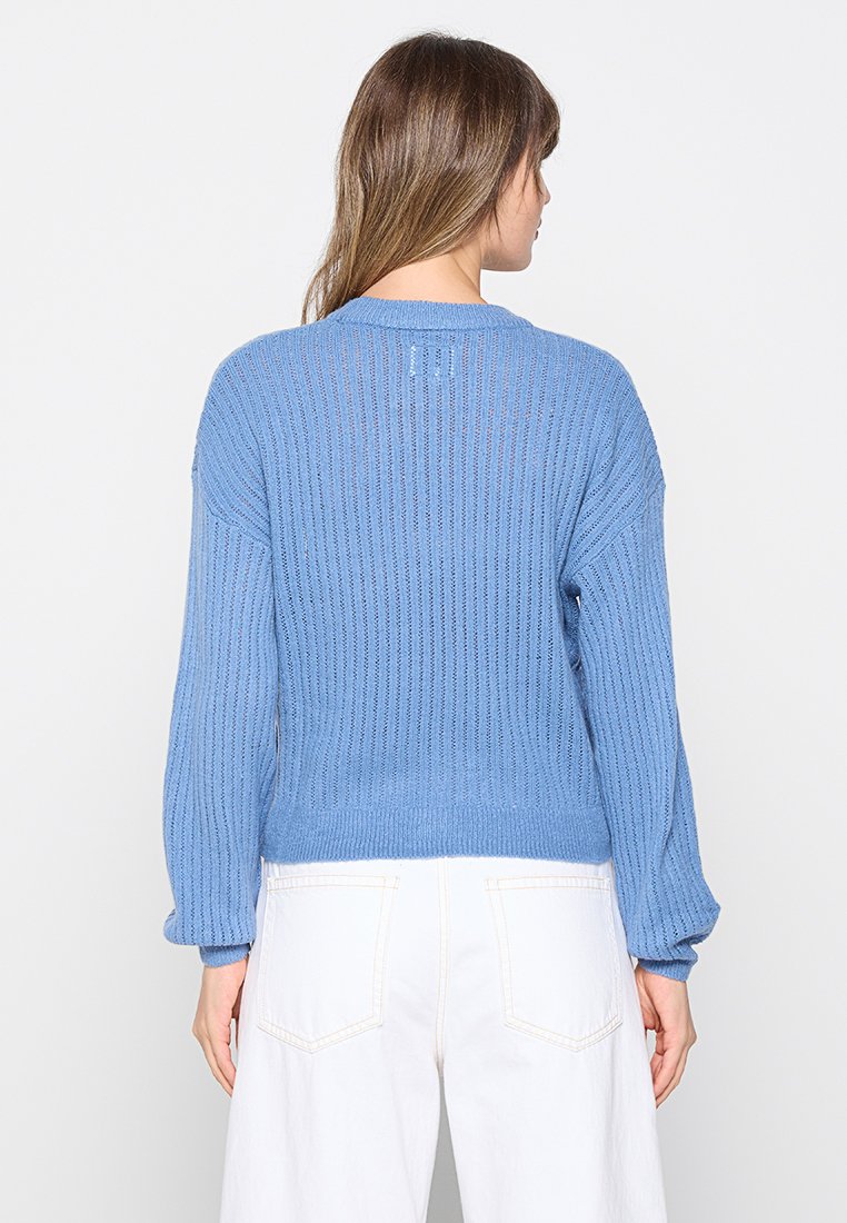 Blauwe gebreide trui met een ribbelstructuur, losse pasvorm en zakmouw. Heeft een ronde halslijn en wordt gedragen over witte broeken.