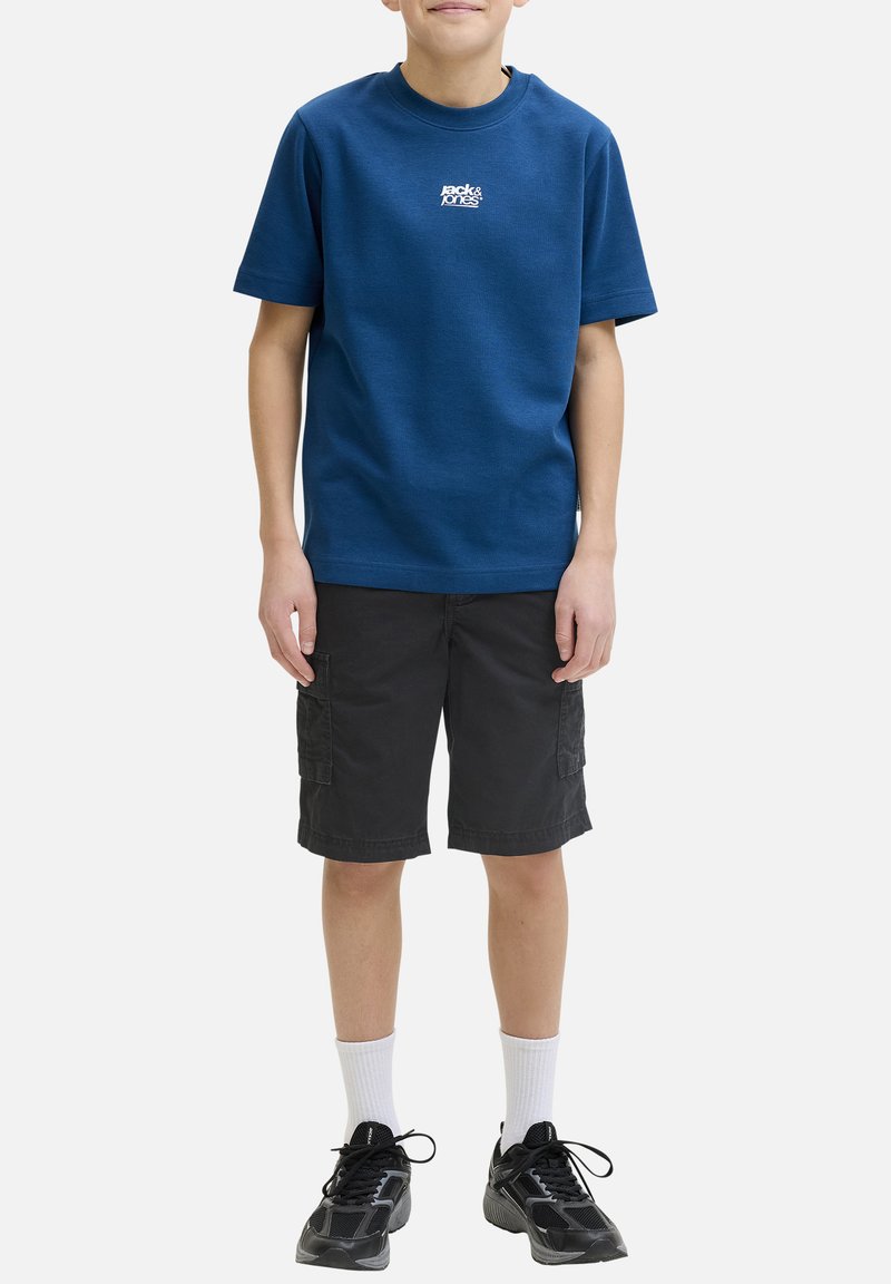 Garçon portant un t-shirt bleu Jack & Jones, un short cargo noir, des chaussettes blanches et des baskets noires, debout les bras détendus le long du corps.