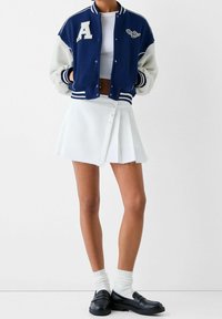 Marinblå och krämfärgad varsityjacka med ett "A"-märke, bärs över en vit crop top och en vit veckad minikjol. Svarta loafers och ribbade strumpor fulländar looken.