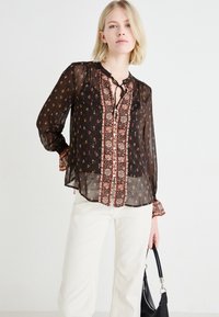 Blusa nera trasparente con motivi floreali e scollo allacciato; presenta maniche lunghe con polsini a volant e una spalla ricamata in contrasto.