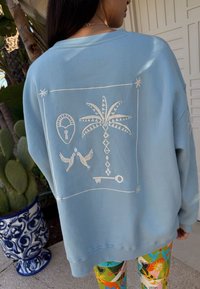 Felpa azzurro chiaro con motivo ricamato bianco sul retro, che presenta una palma, uccelli e forme geometriche. Tessuto morbido, vestibilità comoda.