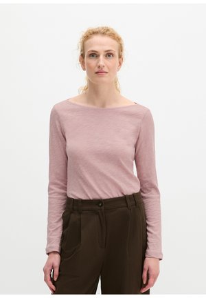 Kobieta z kręconymi blond włosami ubrana w jasnoróżową bluzkę z długim rękawem i ciemnobrązowe spodnie z wysokim stanem, stojąca na jednolitym białym tle.