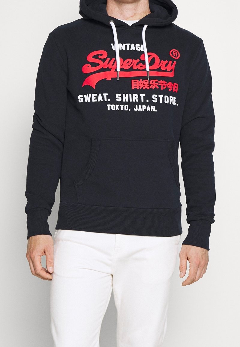 Homme portant un sweat à capuche noir SuperDry avec un texte rouge et blanc, et un pantalon blanc, debout les mains relâchées le long du corps.