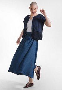 Persona sonriente con cabeza rapada, vestida con blusa blanca, chaqueta azul marino, falda larga azul y zapatos burdeos con cordones, posando con un pie levantado.