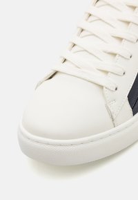 Sneaker bianco in pelle con texture liscia, punta rotonda e lacci piatti, decorata con una striscia blu navy sul lato. Suola in gomma con grip strutturato.