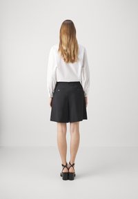 Frau mit langen Haaren, die eine weiße Bluse, schwarze knielange Shorts mit Gesäßtaschen und schwarze Sandalen mit Absatz trägt, steht vor einem einfarbigen Hintergrund.