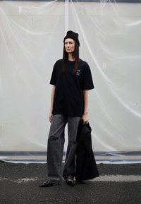 Melnas pārlielas T-krekls, pelēkas norautas plata piegriezuma džinsi, melna cepure un melni norādīti apavi. Tur melnu jaku, stāv uz teksturētas virsmas.