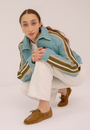 Giacca zip blu chiaro con strisce verdi e beige, abbinata a pantaloni bianchi e scarpe in suede marrone, caratterizzata da un design a scollo basso e lacci.