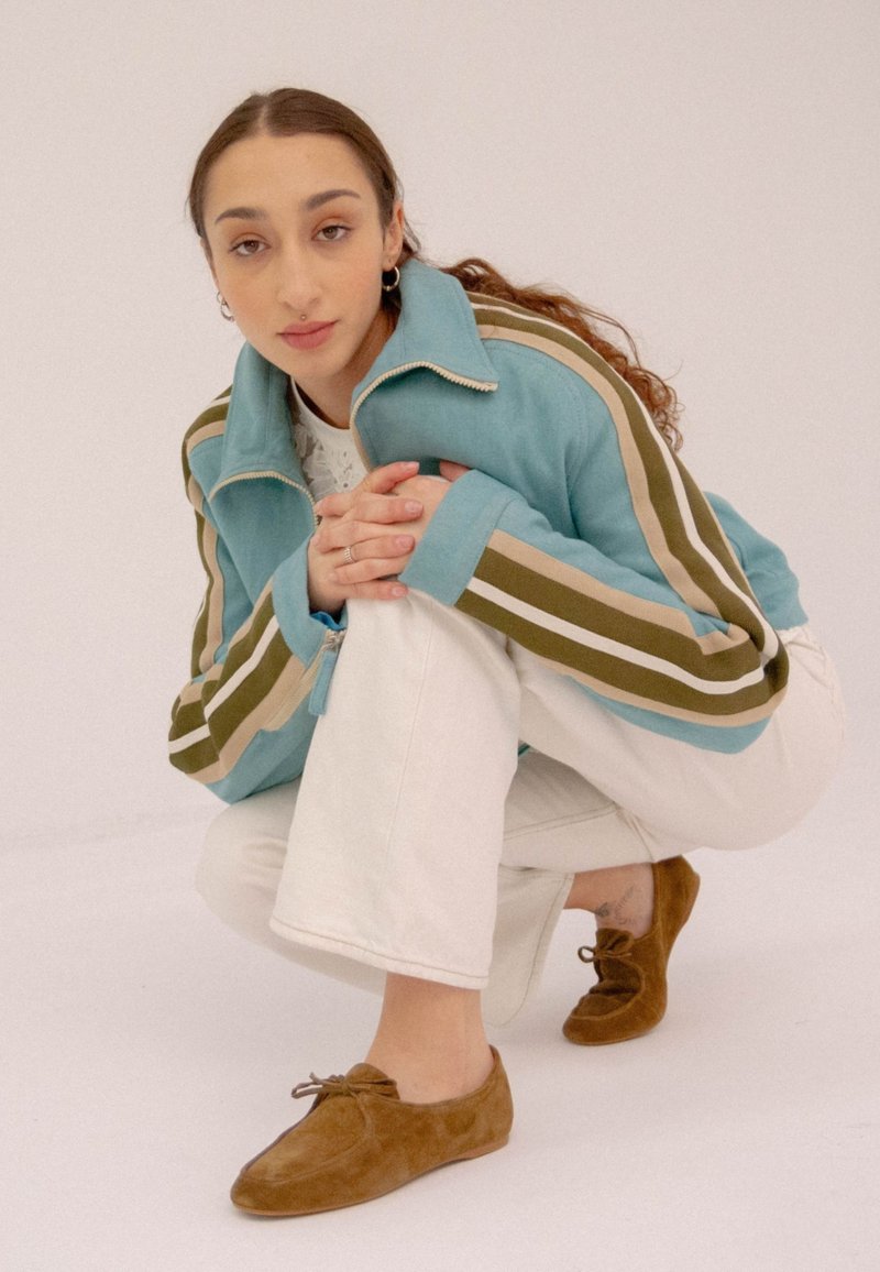 Giacca zip blu chiaro con strisce verdi e beige, abbinata a pantaloni bianchi e scarpe in suede marrone, caratterizzata da un design a scollo basso e lacci.