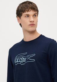 Camiseta de manga larga en azul marino, hecha de algodón, con un contorno blanco del logotipo de un cocodrilo acompañado de texto. Cuello redondo acanalado.