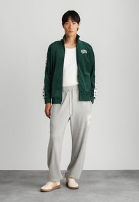 Jaqueta verde de veludo com fecho éclair e detalhes brancos, calças de fato cinzentas e ténis bege. O conjunto apresenta um corte descontraído e estilo casual.